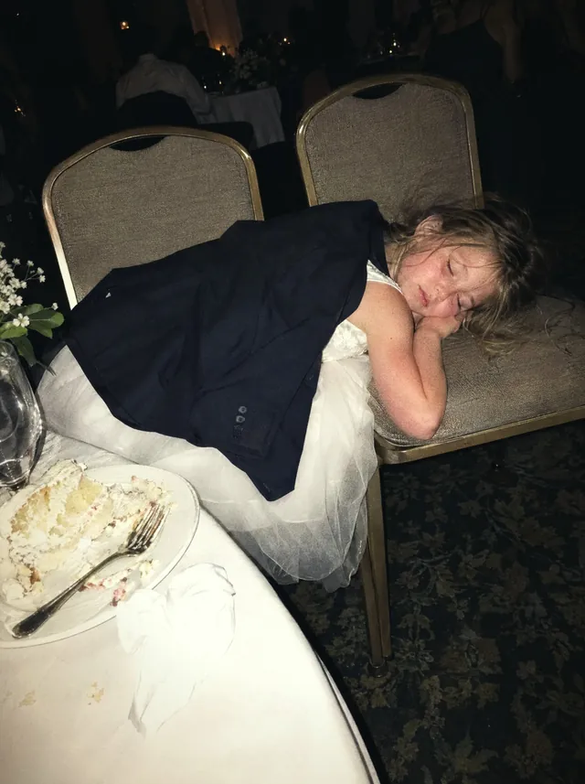 The flower girl asleep across two banquet chairs under a suit jacket, next to a half-eaten slice of cake.