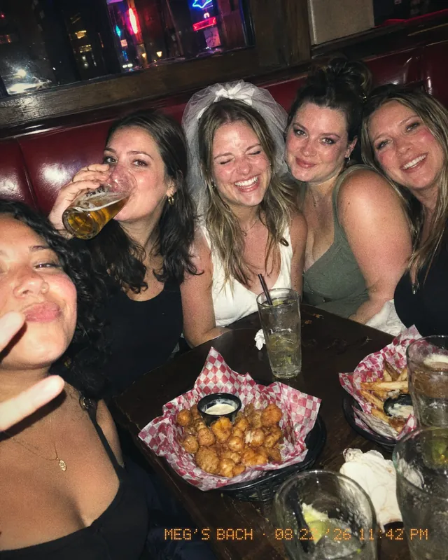 Five friends squeezed into a bar booth, the bride in a veil laughing, baskets of cheese curds on the table.