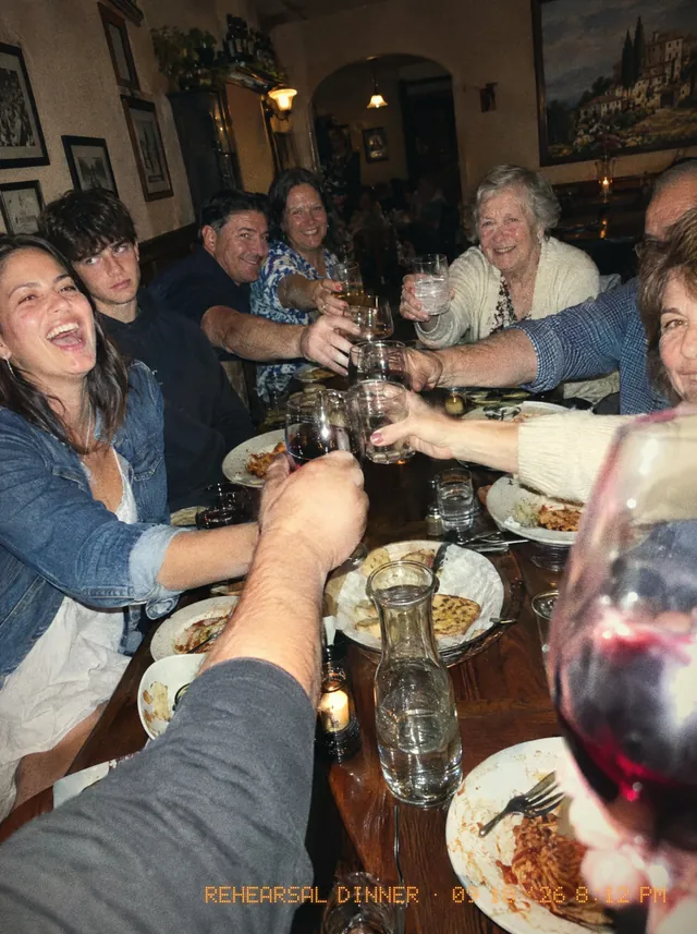 Everyone at a long restaurant table leaning in to clink glasses over plates of pasta, the bride-to-be laughing.