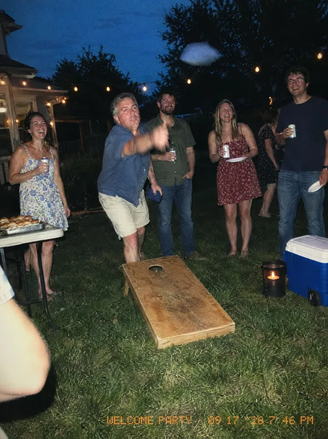 A backyard welcome party at dusk: an uncle mid-throw at bean bags while friends with cans and paper plates laugh under string lights.