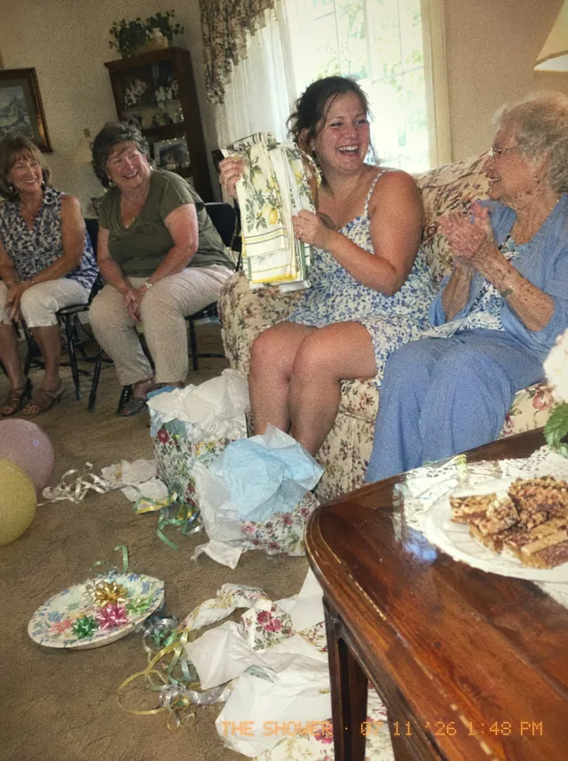 A living-room shower: the bride-to-be on a floral couch holding up kitchen towels, her grandmother clapping beside her, wrapping paper all over the carpet.